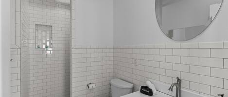 Une salle de bain moderne avec des carreaux blancs, une douche, des toilettes, un lavabo avec un miroir et une trousse de maquillage posée sur le comptoir.