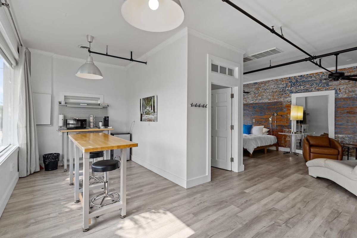 A modern apartment with a kitchen, dining area, and living room. It features a wooden floor, white walls, and a brick accent wall.