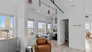 A spacious living area with large windows, a leather armchair, a kitchen area, and a view of the cityscape. - Guest House Topsail (Holly Ridge)