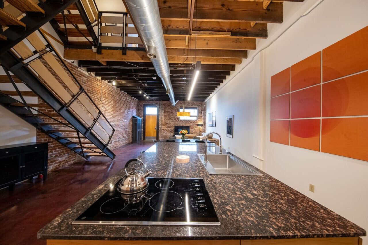 Una cocina moderna con encimera de granito, electrodomésticos de acero inoxidable, paredes de ladrillo visto y una escalera que sube a un nivel superior.