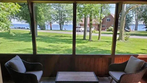 Une pièce dotée d’une grande fenêtre donnant sur un espace verdoyant et un bâtiment situé de l’autre côté de l’eau. On y trouve un canapé en osier avec des coussins et une petite table au centre.