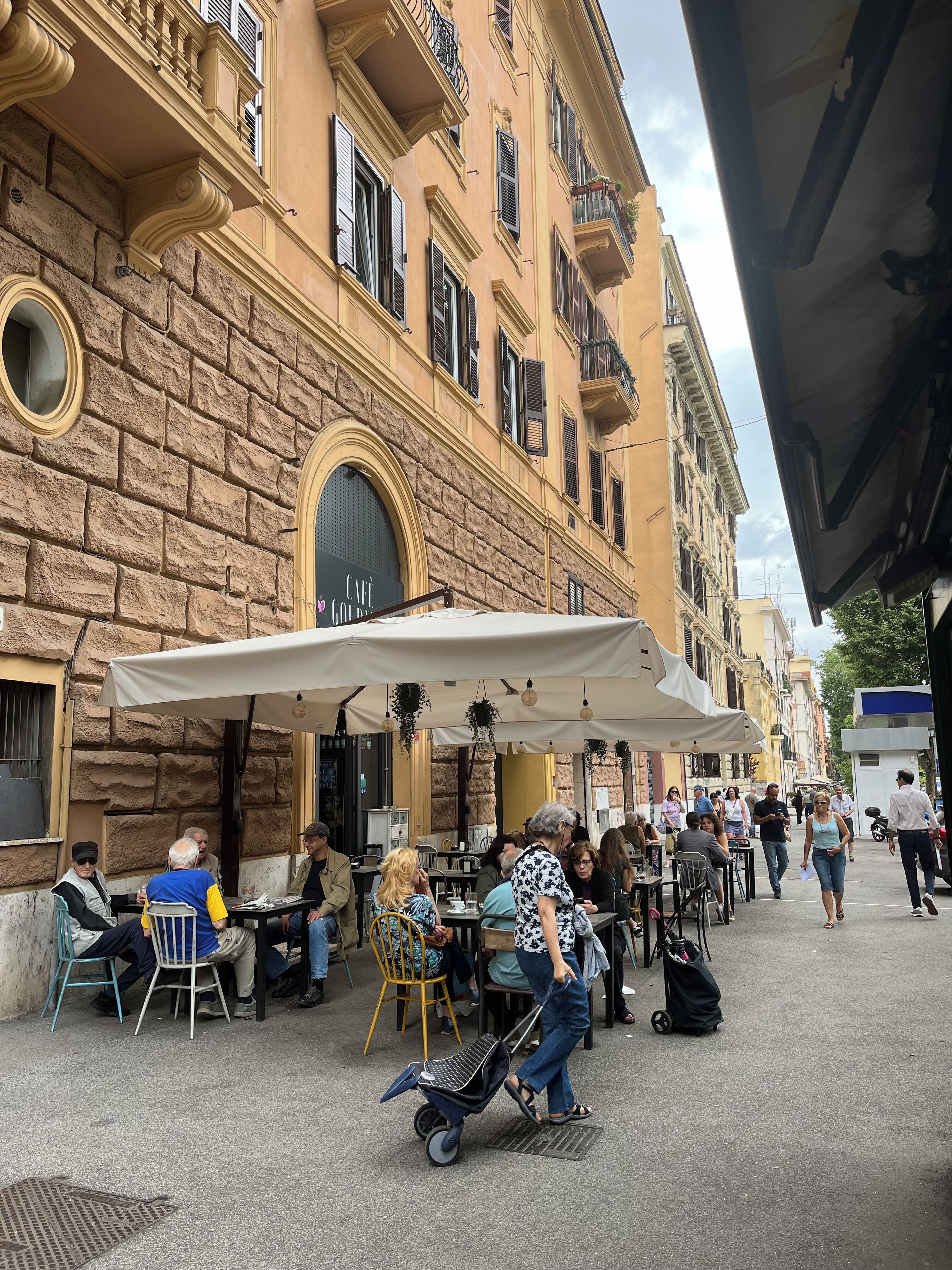 An outdoor cafe with tables and chairs, a stone building with a cafe sign, and a person pushing a stroller.