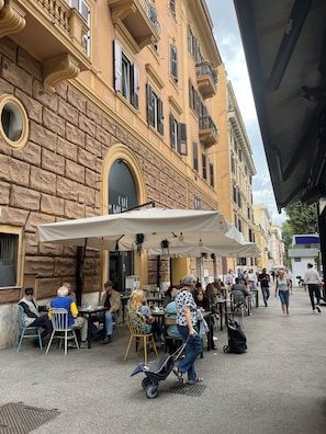 Een terras met tafels en stoelen, een stenen gebouw met een cafébord, en iemand die een kinderwagen duwt.