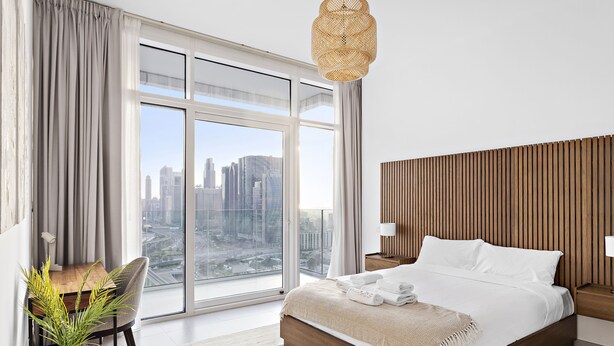 A modern hotel room with a large window, a wooden headboard, a bed with white linens, and a small table with a plant.