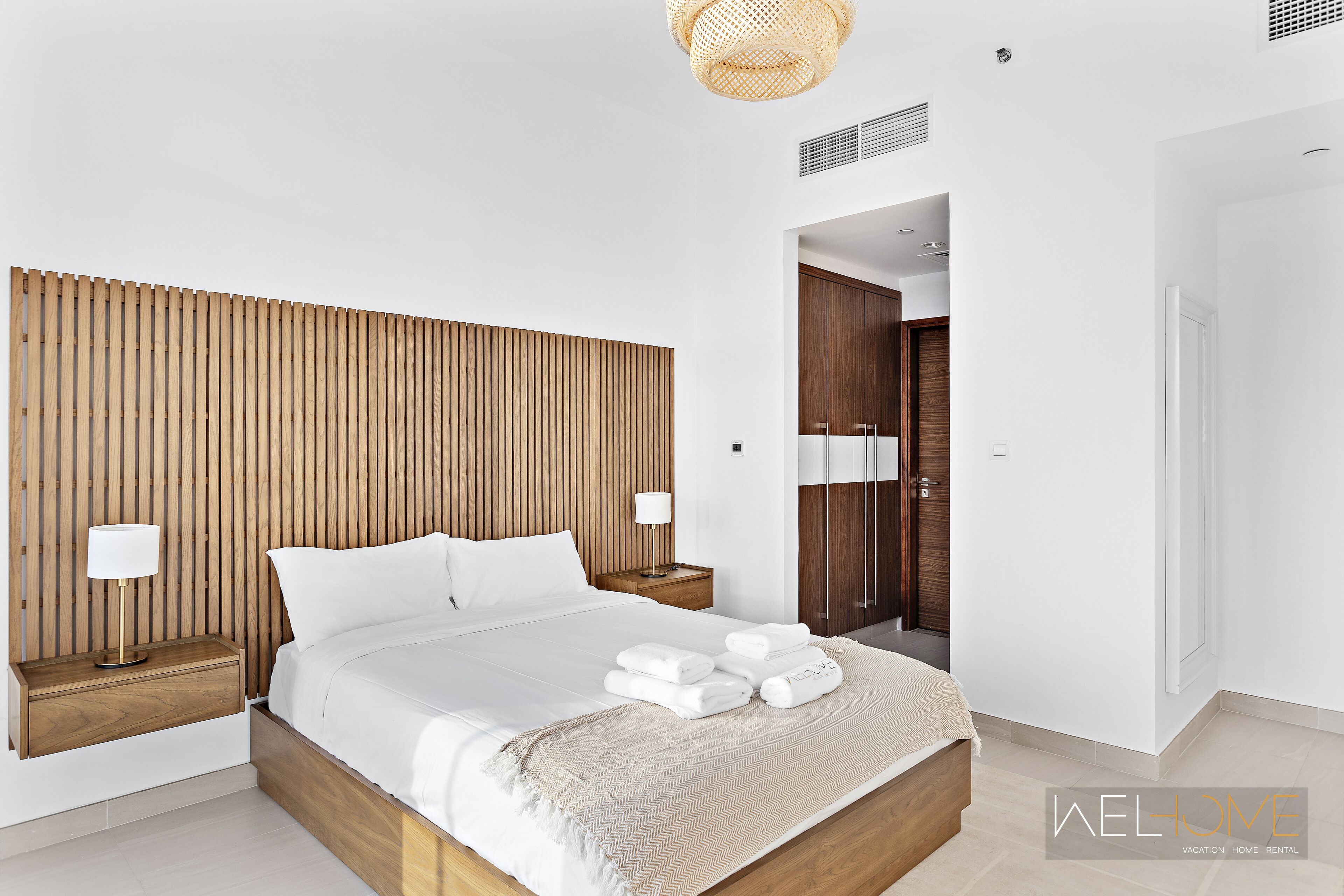 Ein modernes Hotelzimmer mit einem hölzernen Kopfteil, einem ordentlich bezogenen Bett mit weißen Bettwäsche, zwei Nachttischen mit Lampen und einer sichtbaren Tür zu einem weiteren Raum.