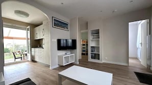 A spacious living area with a flat-screen TV, a dining table, and a kitchen area with cabinets and a window. - Maison Proche de Brest Arena (Brest)