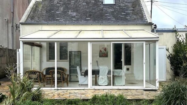 A single-story house with a glass conservatory. Inside, there is a dining table with chairs and a white chair. - Maison Proche de Brest Arena (Brest)