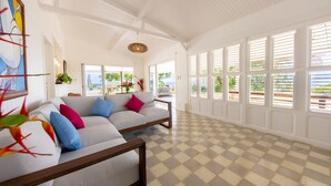 Un salón espacioso con piso de cuadros, un sofá blanco con cojines coloridos y ventanas grandes con contraventanas.