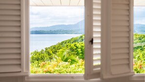 Vista a través de una ventana abierta con contraventanas blancas, mostrando un paisaje exuberante con vegetación y un cuerpo de agua.