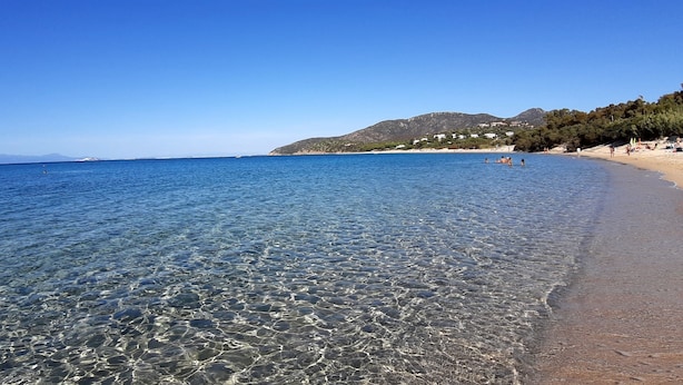 Uma praia arenosa com águas calmas e pedras visíveis, com montanhas ao fundo.