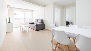Een lichte, moderne woonkamer met een grijze bank, een witte salontafel, en een eethoek met een witte tafel en stoelen.