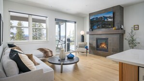 Una sala de estar moderna con chimenea, un sofá, un comedor y un amplio ventanal con vistas a árboles y montañas.