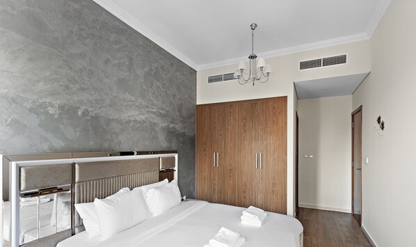 A modern hotel room with a large bed, a wooden wardrobe, a built-in headboard, and a chandelier.