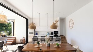 Una cocina moderna con armarios blancos, una mesa de comedor de madera y sillas negras. Hay lámparas de mimbre colgantes y un reloj en la pared.