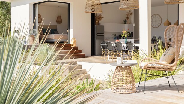 Un patio moderno al aire libre con mobiliario de mimbre, una mesa de comedor y una cocina visible a través de un arco abierto.