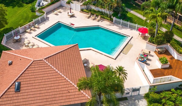 An aerial view of a swimming pool with a tiled roof, surrounded by lounge chairs and umbrellas, with palm trees and a well-maintained lawn.