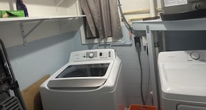 A laundry area with two washing machines, a window with curtains, and a shelf with cleaning supplies.