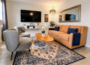 A living room with a sofa, two armchairs, a coffee table, a TV, a wall-mounted mirror, a hanging light fixture, and a patterned rug.