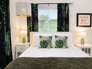 A bedroom with a bed, two bedside tables, a lamp, a window with curtains, and a framed picture on the wall.