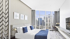 A modern hotel room with a large bed, a desk, and a view of the cityscape through large windows.