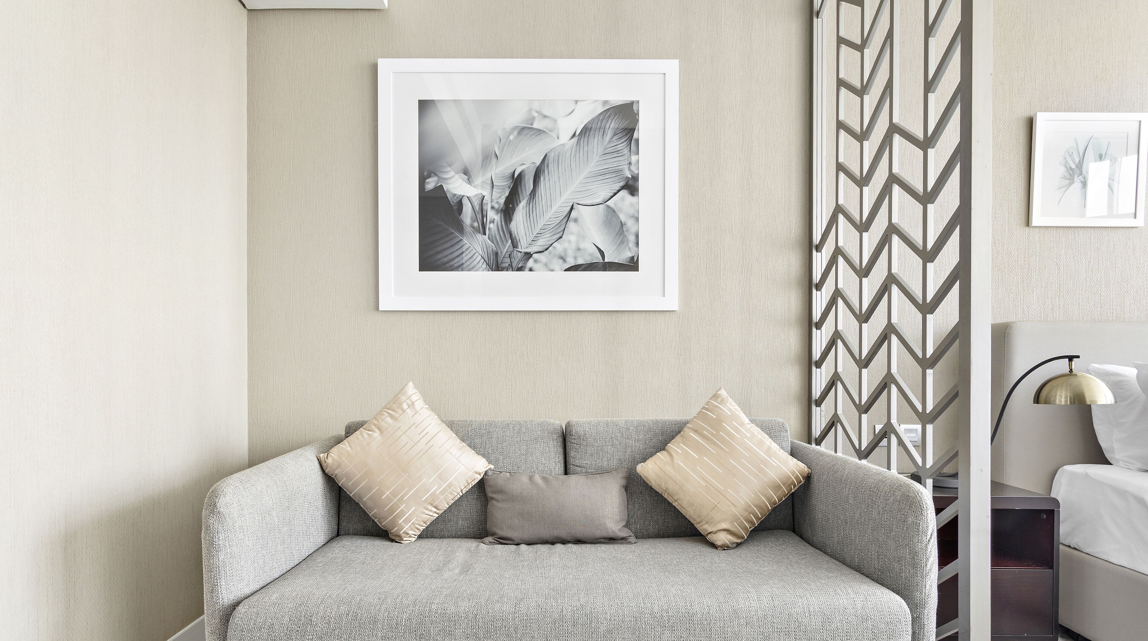 Un salon aux couleurs neutres, avec un canapé gris, deux coussins, une photographie en noir et blanc encadrée, un mur à motifs et un lit avec une tête de lit.