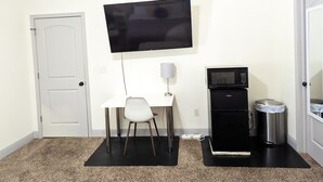 Ein kleines Zimmer mit einer weißen Tür, einem weißen Schreibtisch mit Stuhl, einem fest montierten Flachbildfernseher, einer Mikrowelle und einem Mülleimer.