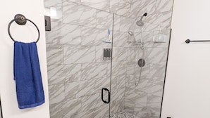Ein modernes Badezimmer mit einer Glaskabine zur Dusche, Marmorfliesen, einem blauen Handtuch und einem Mülleimer.