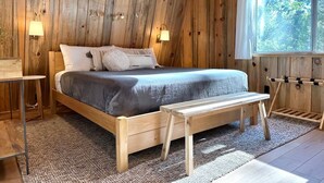 Dormitorio con paneles de madera, una cama, lámparas de noche, un banco y una ventana con vistas a árboles.