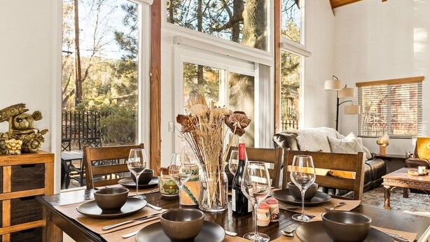 A dining area with a wooden table set for four, surrounded by large windows offering a view of trees and a forest.