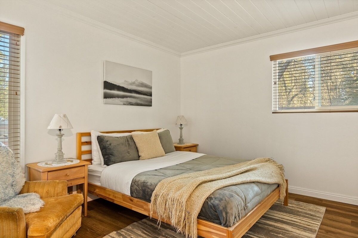 Un dormitorio con una cama de madera, una mesita de noche con lámpara, una ventana con persianas y un cuadro en la pared.
