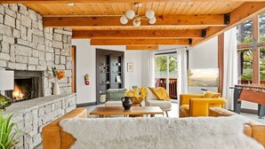 A cozy living room with a stone fireplace, wooden ceiling beams, and a large window offering a scenic view. - Heart of Big Bear | Lake & Mountain View (Big Bear Lake)