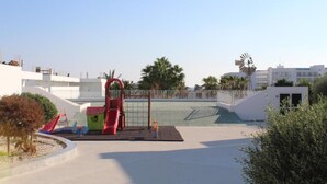 Ein Spielplatz mit einer roten Rutsche und einer grünen Kletterkonstruktion, umgeben von einem weißen Zaun und Palmen.