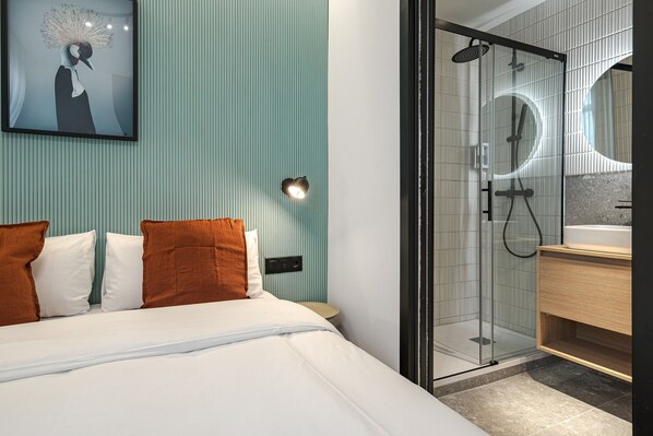 A modern hotel room with a bed, a glass shower enclosure, a round mirror, and a wooden cabinet.