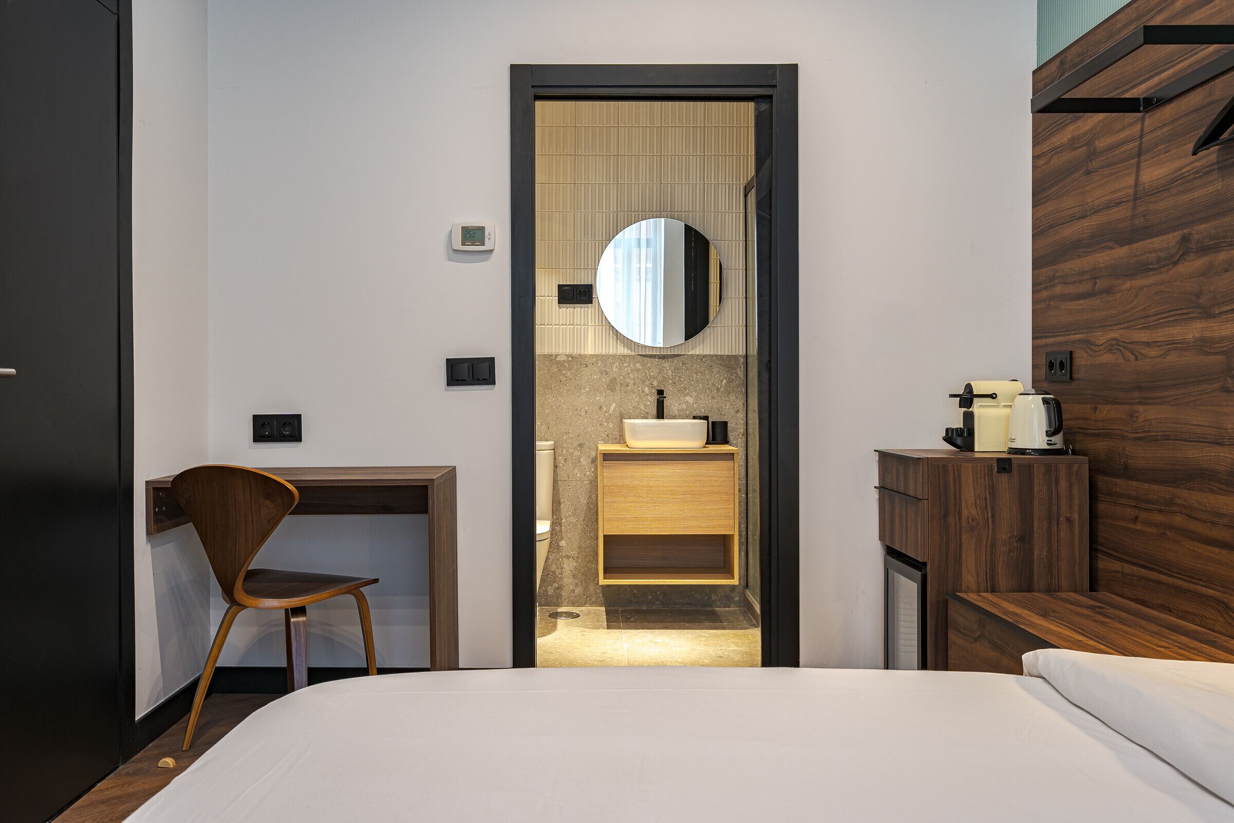 A modern hotel room with a wooden desk, a chair, a bed, and a bathroom with a mirror and sink.