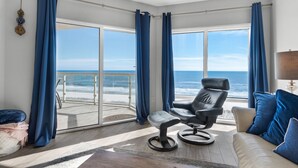 Une pièce dotée d’une grande porte-fenêtre coulissante donnant sur un balcon avec vue sur l’océan. On y trouve un fauteuil en cuir noir et un canapé beige agrémenté de coussins bleus.