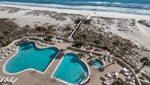 Vue aérienne d’un complexe hôtelier en bord de mer, avec deux piscines, une plage et une jetée.