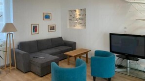 A modern living room with a grey sofa, two blue armchairs, a wooden coffee table, a TV, and artwork on the walls. - Maison Pêcheur Cosy Proche Plage et Port 6pers (Les Sables-d'Olonne)