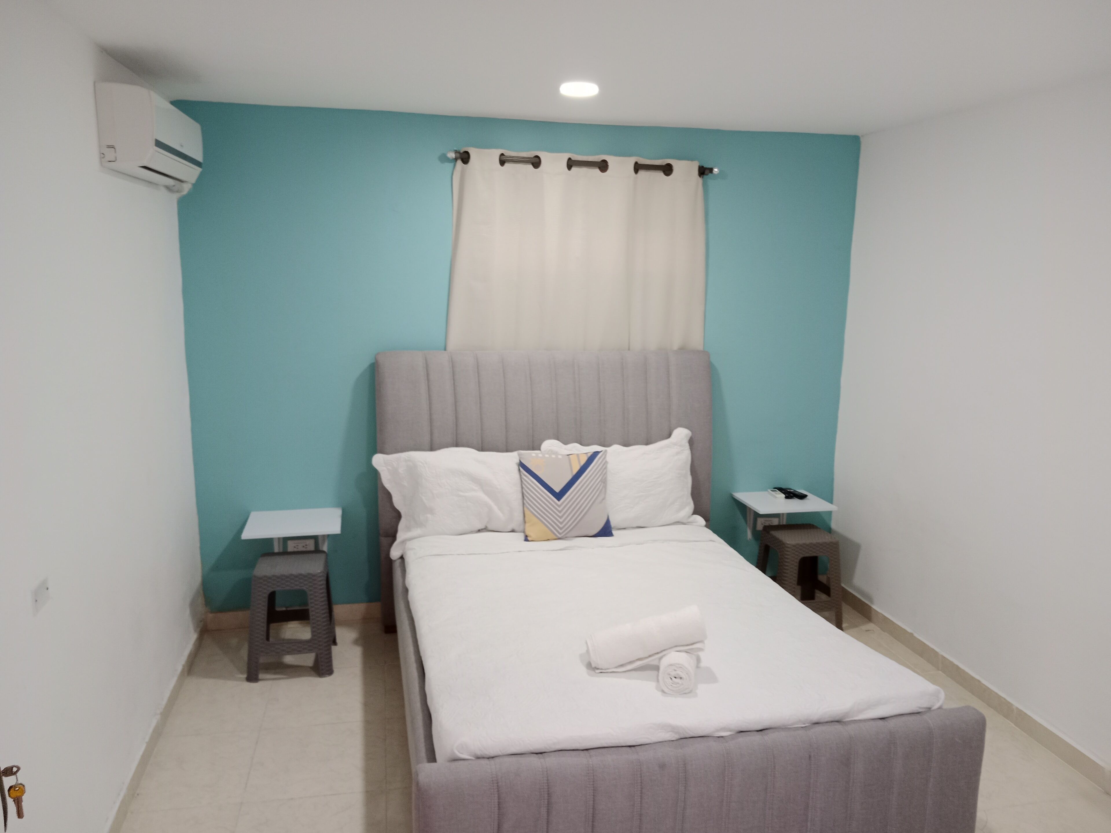 Cama bem arrumada com cabeceira cinza e roupa de cama branca, duas mesas de cabeceira com abajures e uma parede cor de verde-água.