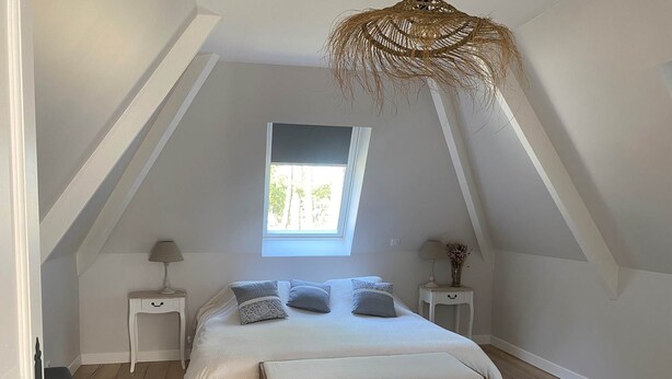 A bedroom with a sloped ceiling, a bed with a wooden headboard, two bedside tables with lamps, a window, and a woven ceiling light fixture.