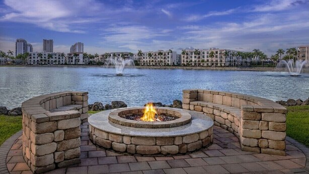 A stone fire pit with seating, overlooking a lake with fountains and buildings in the background. - 2BR Villa at Marriott Villas at Doral – Full Kitchen + Golf & Resort Amenities (Miami)