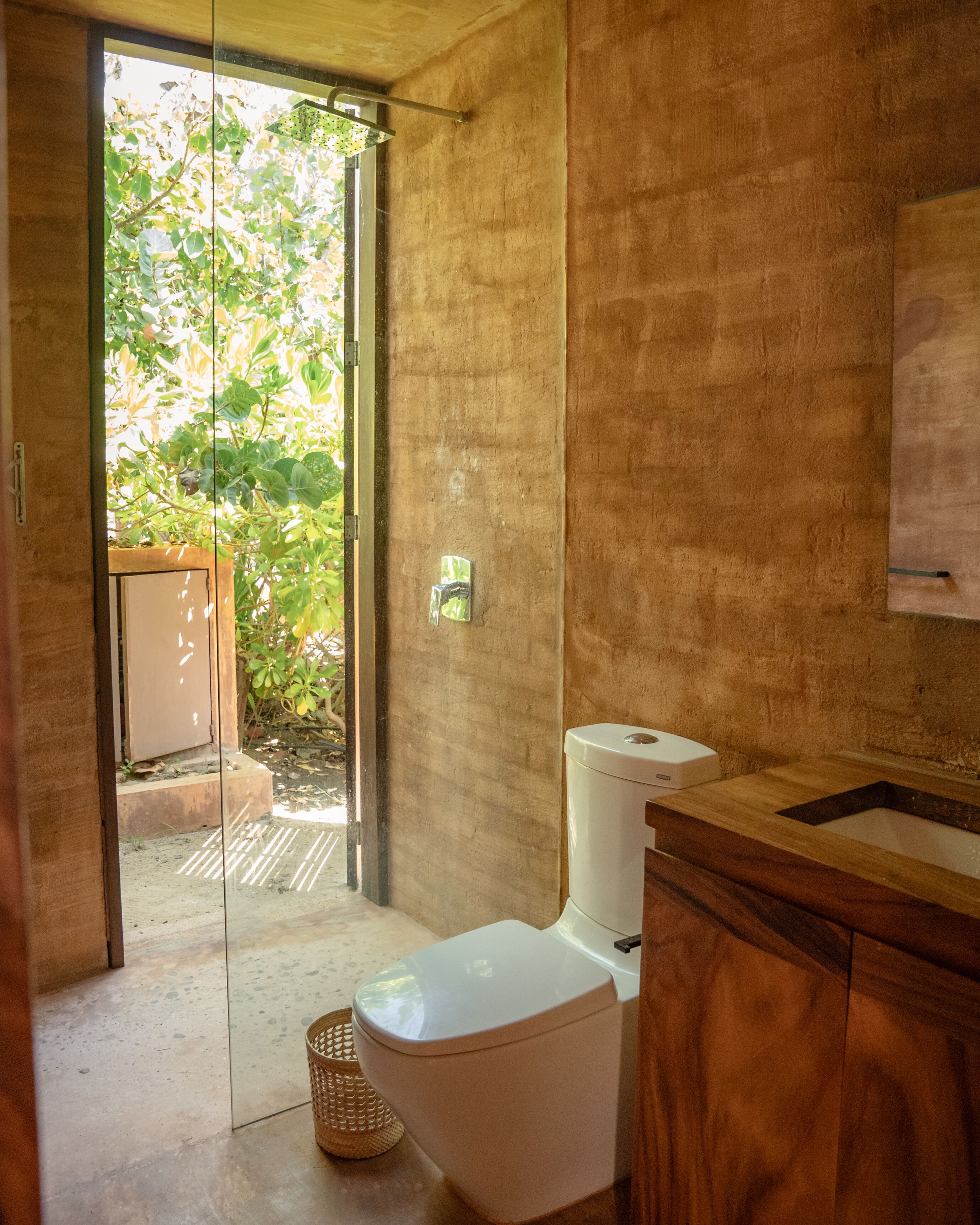 Et badeværelse med toilet, vask og bruseområde. Et vindue giver naturligt lys og udsigt til grøn vegetation udenfor.