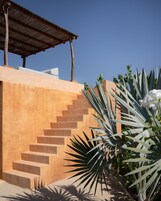 Una escalera conduce a un edificio con techo de madera, rodeado de plantas y un cielo azul despejado.