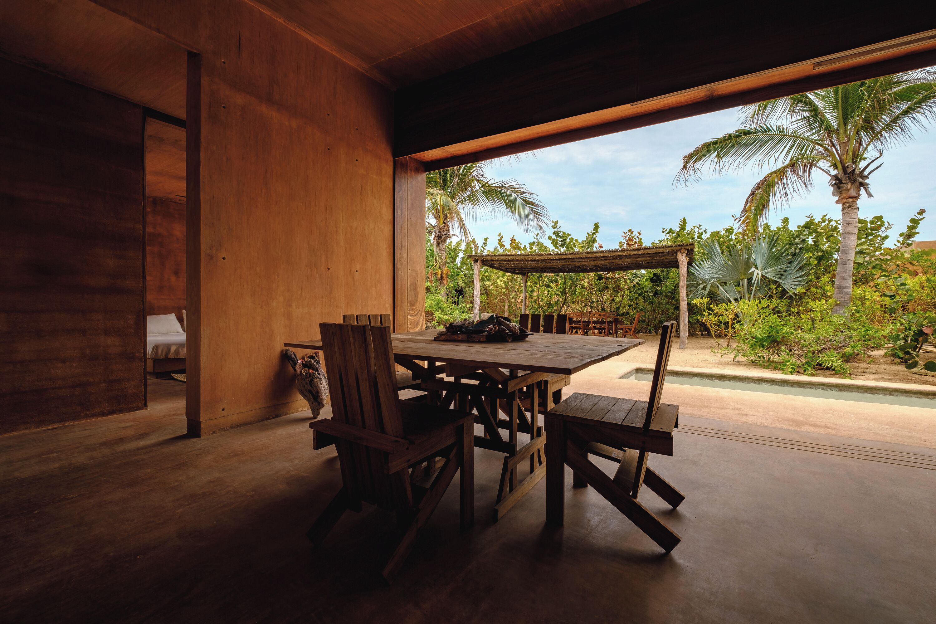 Un comedor de madera con mesa y sillas, un área de estar al aire libre cubierta, y vistas a la vegetación tropical.