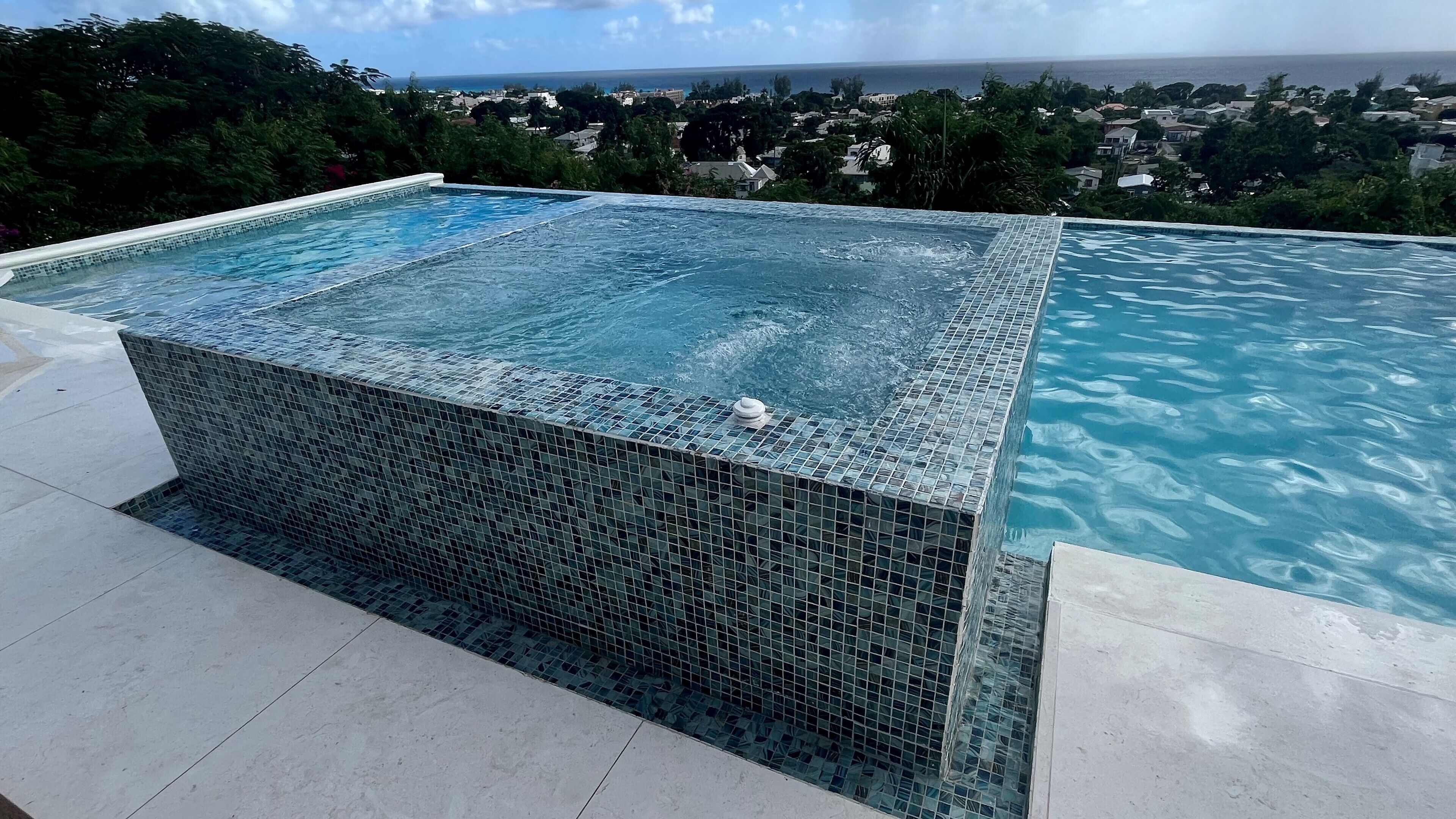 Piscina com área de hidromassagem revestida e vista para o oceano e a cidade ao fundo.