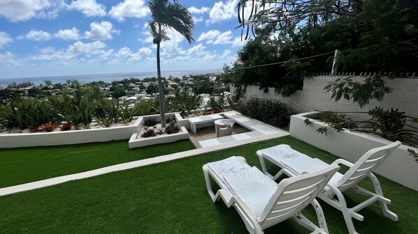 A patio with white lounge chairs and a small table, surrounded by green artificial grass and various plants.
