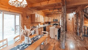 A wooden cabin interior with a dining area, kitchen, and large windows. - HotTub-PoolTbl-Close to Caves-Outdoor TV/Fireplace (Logan)