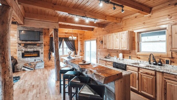 A log cabin interior with a stone fireplace, wooden bar, and a kitchen with wooden cabinets and a large window. - HotTub-PoolTbl-Close to Caves-Outdoor TV/Fireplace (Logan)