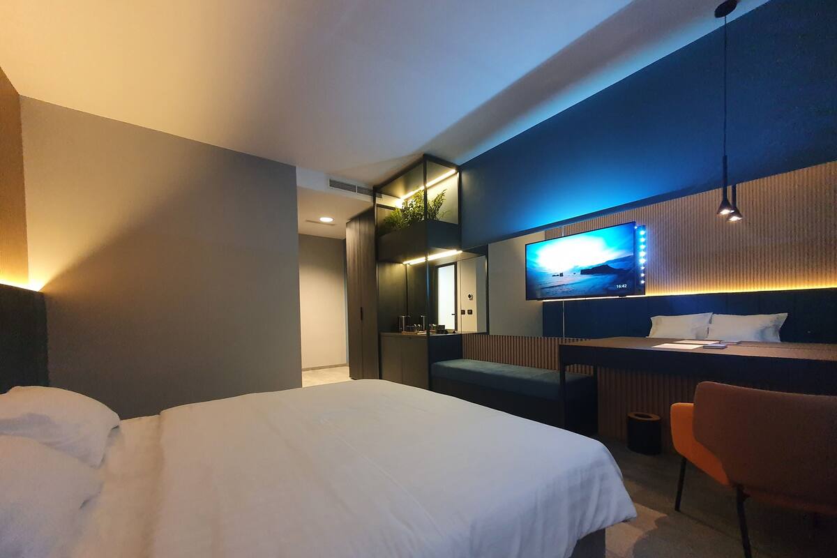Habitación de hotel moderna con una cama grande, un escritorio con silla, televisión montada en la pared y un pequeño rincón de descanso.