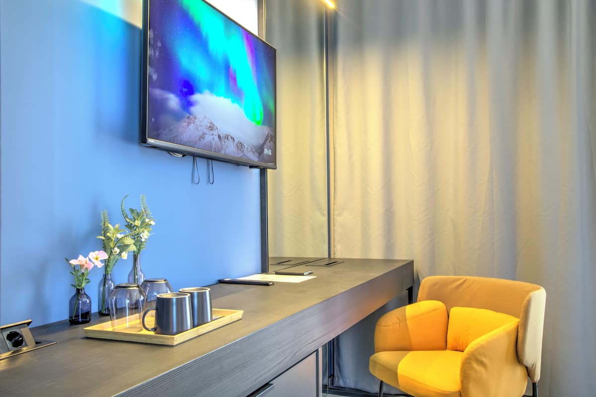 Habitación de hotel moderna con una amplia televisión de pantalla plana mostrando una colorida aurora boreal, un escritorio de madera con una bandeja que contiene tazas y un bloc de notas, y una silla de color amarillo brillante.