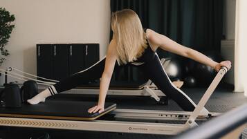 Een persoon doet een rekoefening op een Pilates reformer in een ruimte met een donker gordijn, een wandplank met spullen en een potplant.